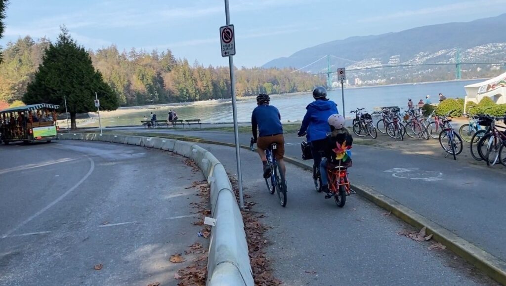 A pair of cyclists riding in the separated and protected bike lane on Stanley Park Drive. One of the two is on a cargo bike with a child in the back seat.